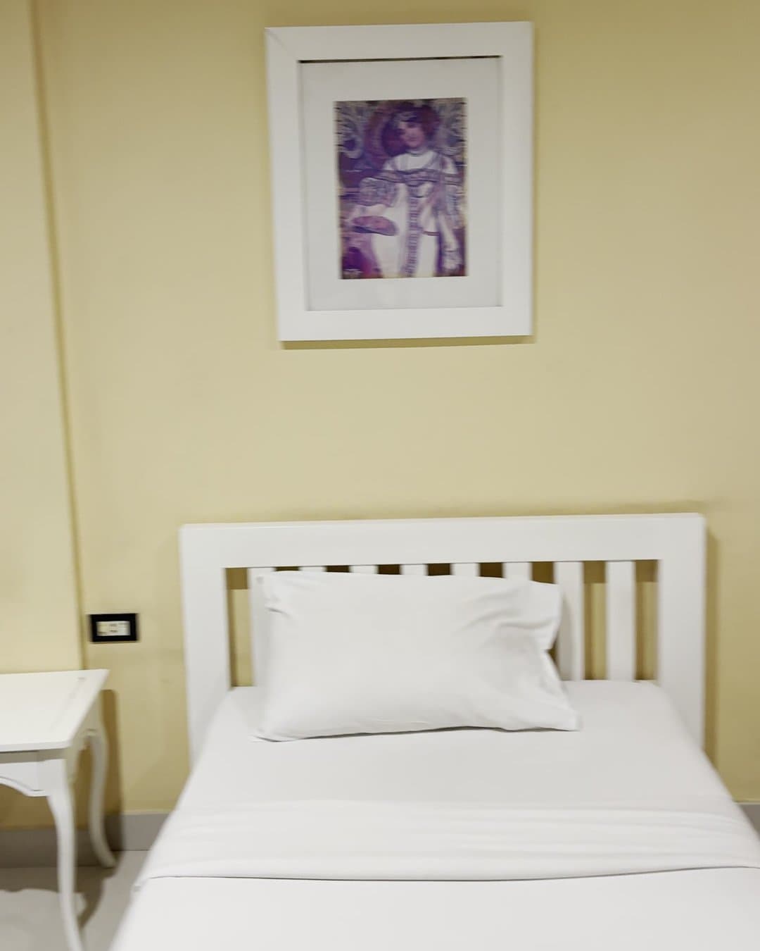 Single bed with a white wooden frame and white linen against a yellow wall with a framed print